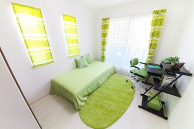 A standard bedroom showcasing a desk, a chair and a single bed. thee curtains, bed sheets, and a floor mat are green colour.